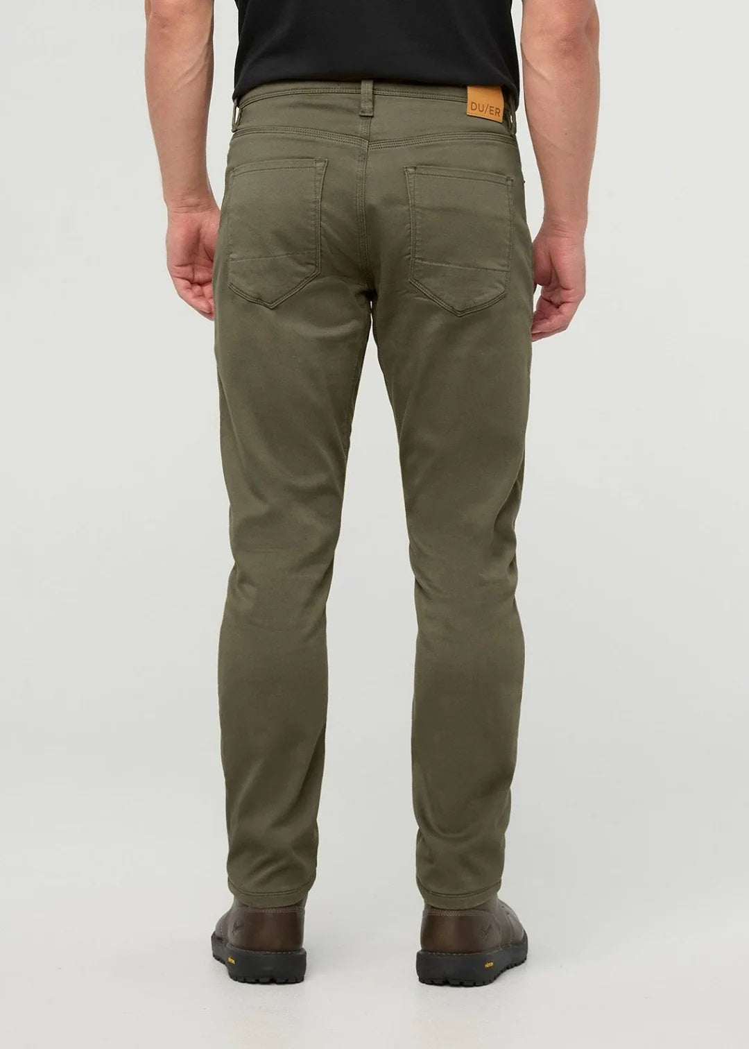 A person stands facing away from the camera against a plain background, wearing DUER - NO SWEAT ATHLETIC TAPER PANTS IN KELP paired with brown shoes.