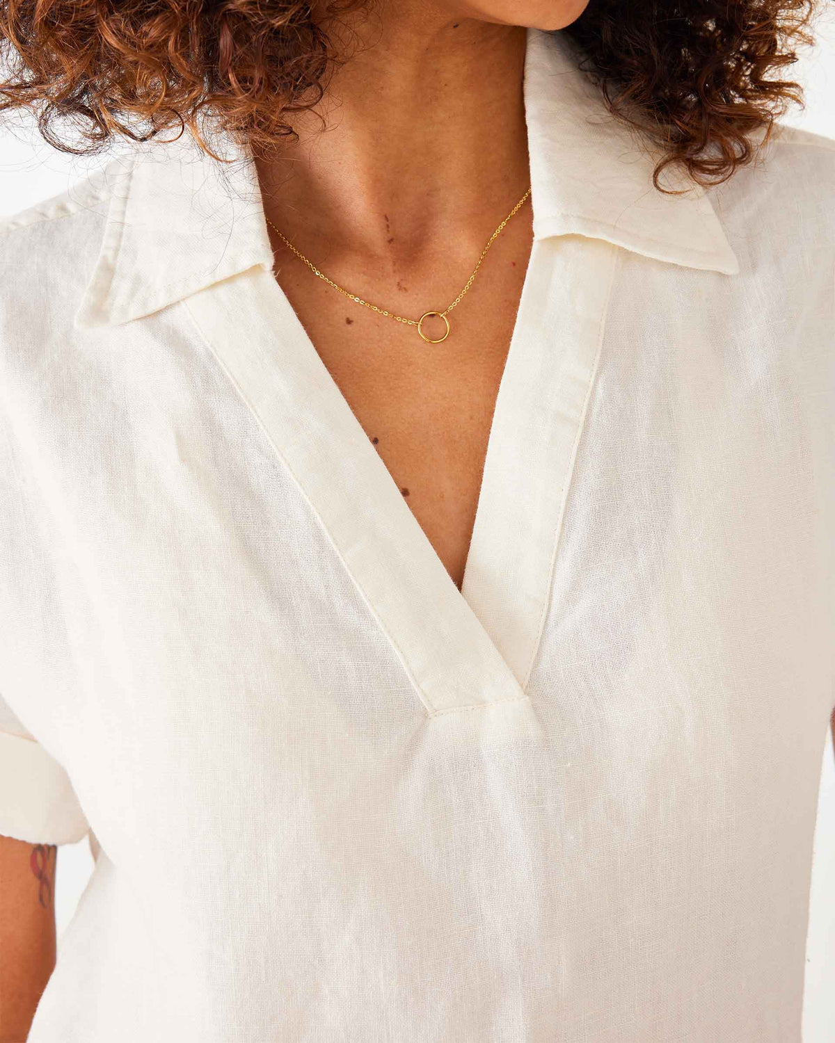 A woman with curly hair wears sunglasses and the MERSEA HADLEY BOXY LINEN SHIRT in cream, featuring loose short sleeves, paired with beige high-waisted pants with front pockets, standing against a white background.