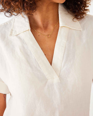 A woman with curly hair wears sunglasses and the MERSEA HADLEY BOXY LINEN SHIRT in cream, featuring loose short sleeves, paired with beige high-waisted pants with front pockets, standing against a white background.
