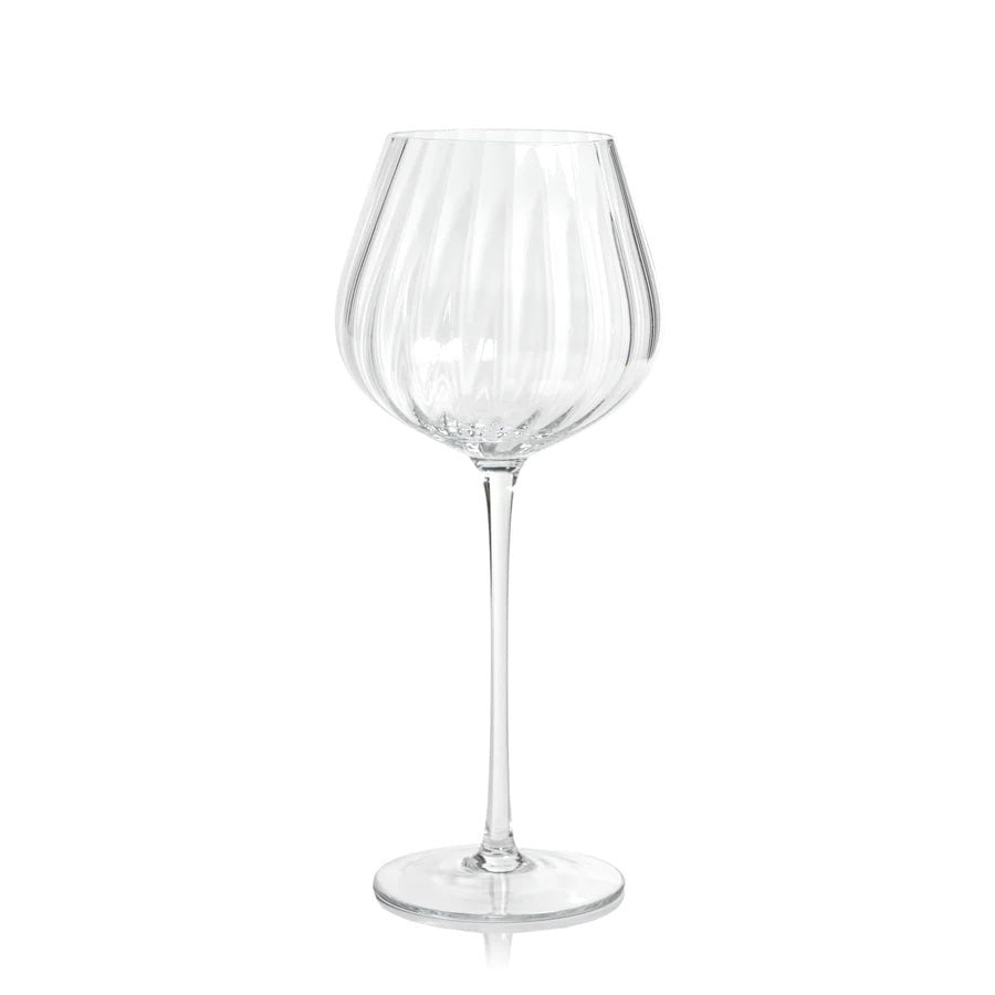A MADELEINE OPTIC RED WINE GLASS by ZODAX stands on a white background. This elegant glass, with a capacity of 580 ml, features a long, slender stem and a wide, slightly curved bowl adorned with vertical ribbed detailing. Its round, flat base ensures stability for the delicate glassware.