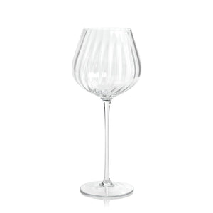 A MADELEINE OPTIC RED WINE GLASS by ZODAX stands on a white background. This elegant glass, with a capacity of 580 ml, features a long, slender stem and a wide, slightly curved bowl adorned with vertical ribbed detailing. Its round, flat base ensures stability for the delicate glassware.