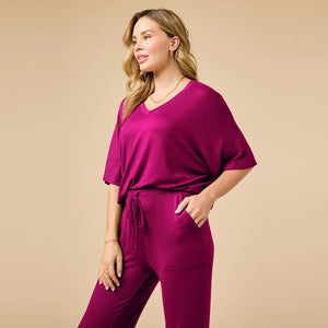 A woman wears a SOFTIES - DREAM RELAXED V-NECK WITH CAPRI LOUNGE SET in magenta, featuring a V-neck top with elbow-length sleeves and matching cropped drawstring pants, paired with cream-colored loafers.