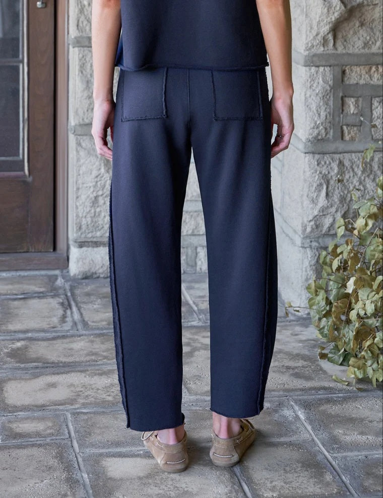 A woman with wavy brown hair smiles as she stands in a bright room, wearing the FRANK & EILEEN Bibi Barrel Leg Sweatpant in Classic Navy paired with a matching short-sleeve top and tan loafers.
