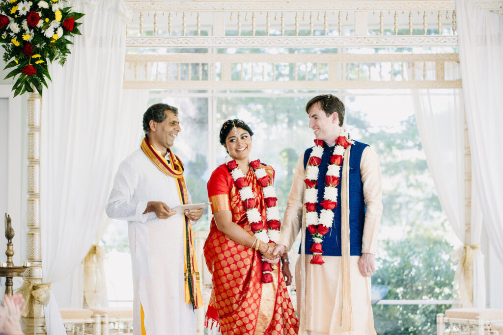 A smiling couple dressed in traditional Indian wedding attire stands together holding hands, adorned with red and white flower garlands, next to a priest in white robes under a decorated mandap.