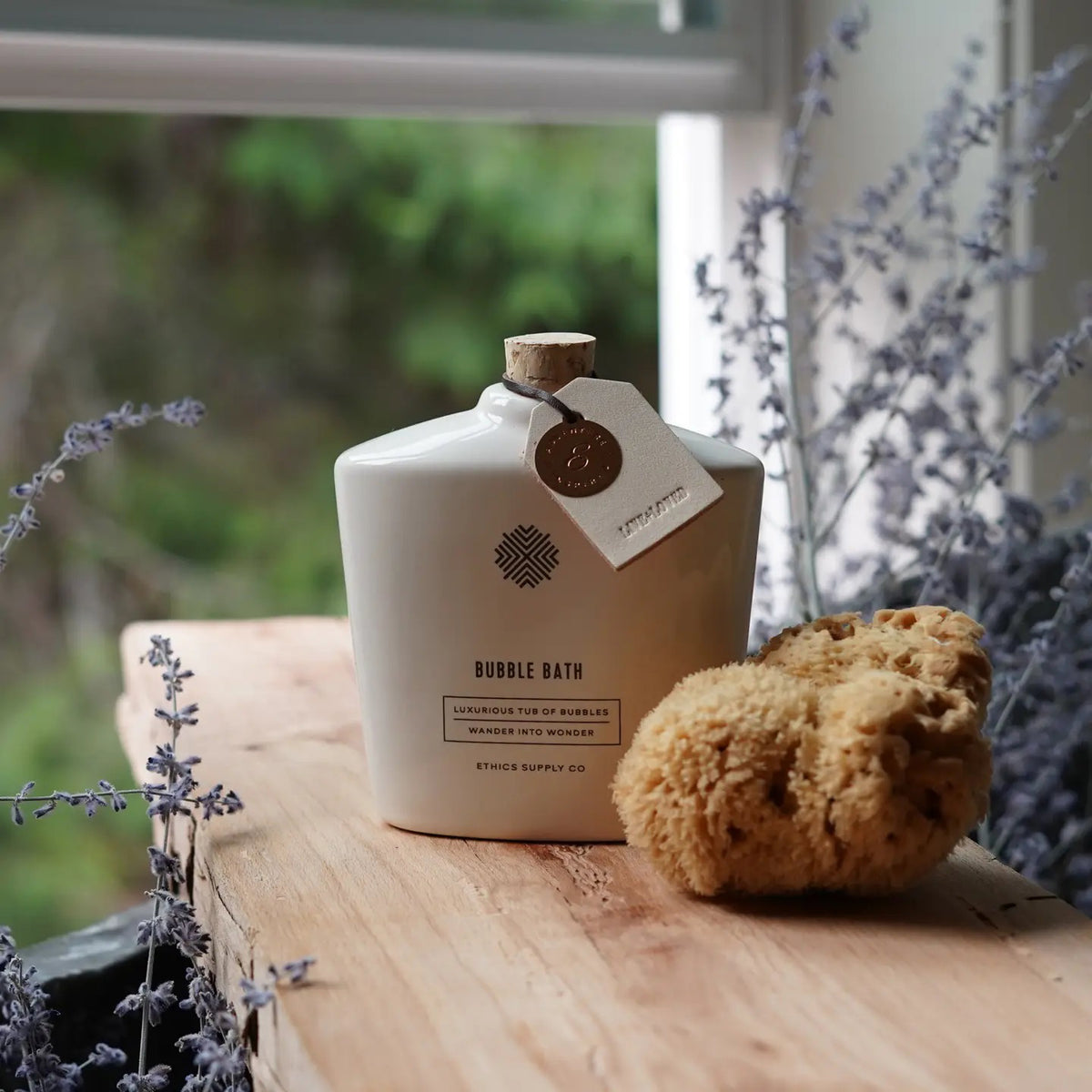 A vegan bubble bath in a luxurious white bottle from Ethics Supply Co features a cork stopper and tag. The label, "Soak In Love Ceramic Bubble Bath," offers an enchanting experience with its promise of abundant bubbles.