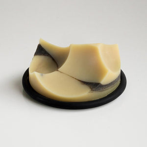 A bar of handmade soap with geometric curves and beige, cream, and dark gray layers rests on the UME STUDIO UME - BLACK BOUTON DISH, displayed against a plain white background.