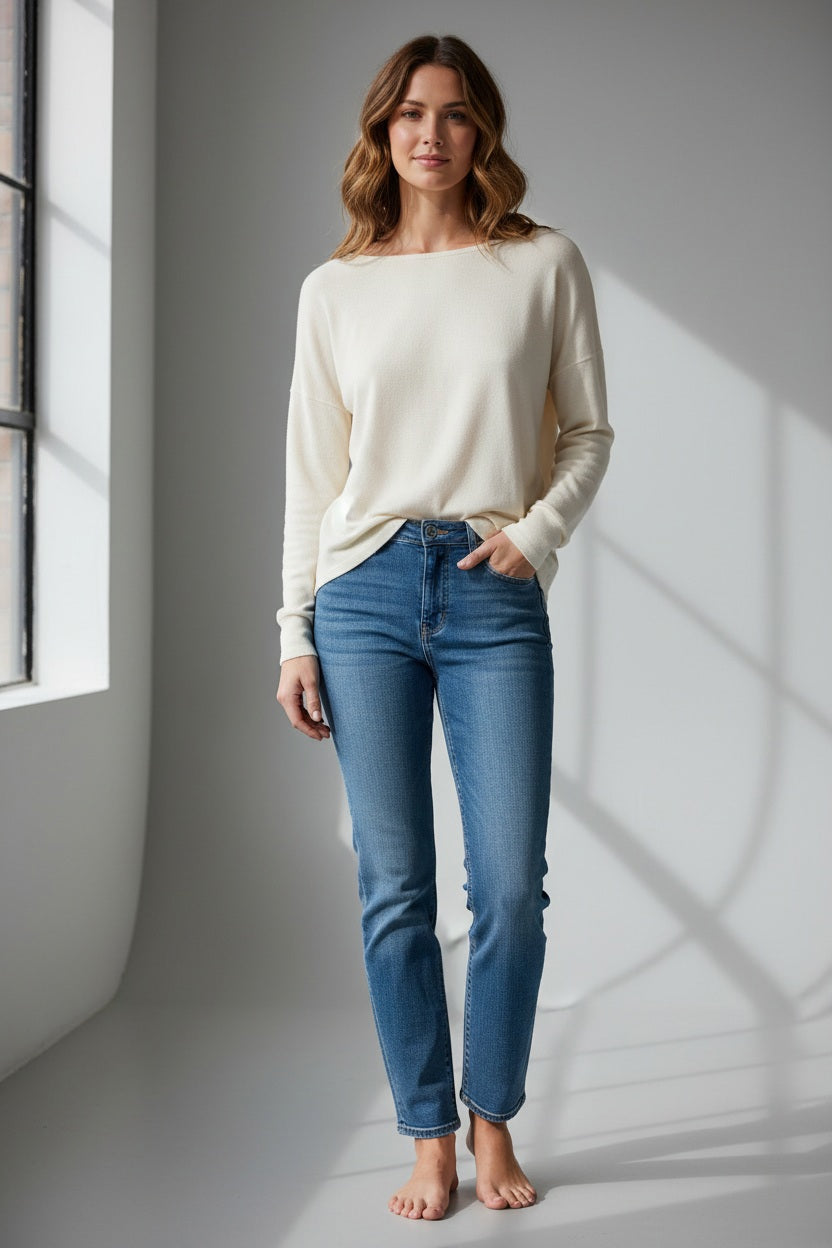 A person with long blonde hair, dressed in the FRANK &amp; EILEEN ELLA Long Sleeve Boatneck Tee in Vintage White from the California Heritage Collection and blue jeans, stands outdoors on a sunny day, glancing over their shoulder. A wooden fence and trees provide a scenic background.