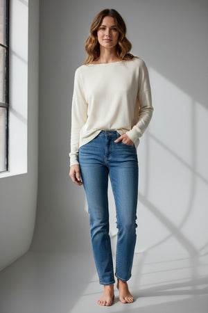 A person with long blonde hair, dressed in the FRANK & EILEEN ELLA Long Sleeve Boatneck Tee in Vintage White from the California Heritage Collection and blue jeans, stands outdoors on a sunny day, glancing over their shoulder. A wooden fence and trees provide a scenic background.
