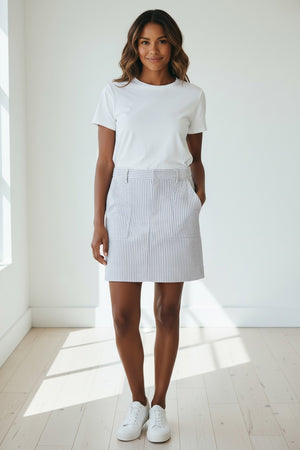 A woman stands outdoors in front of a hedge, barefoot on the grass, wearing a sleeveless white top paired with the FRANK & EILEEN Jameson Skirt in Navy and White Stripe, its utility pockets holding her hands casually.