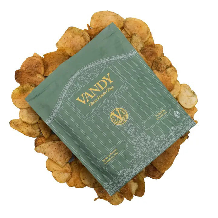 An upright green bag of ANCIENT CRUNCH - VANDY POTATO CHIPS, HERBES DE PROVENCE features gold accents and a gate design, with tallow-cooked potato chips scattered at its base.