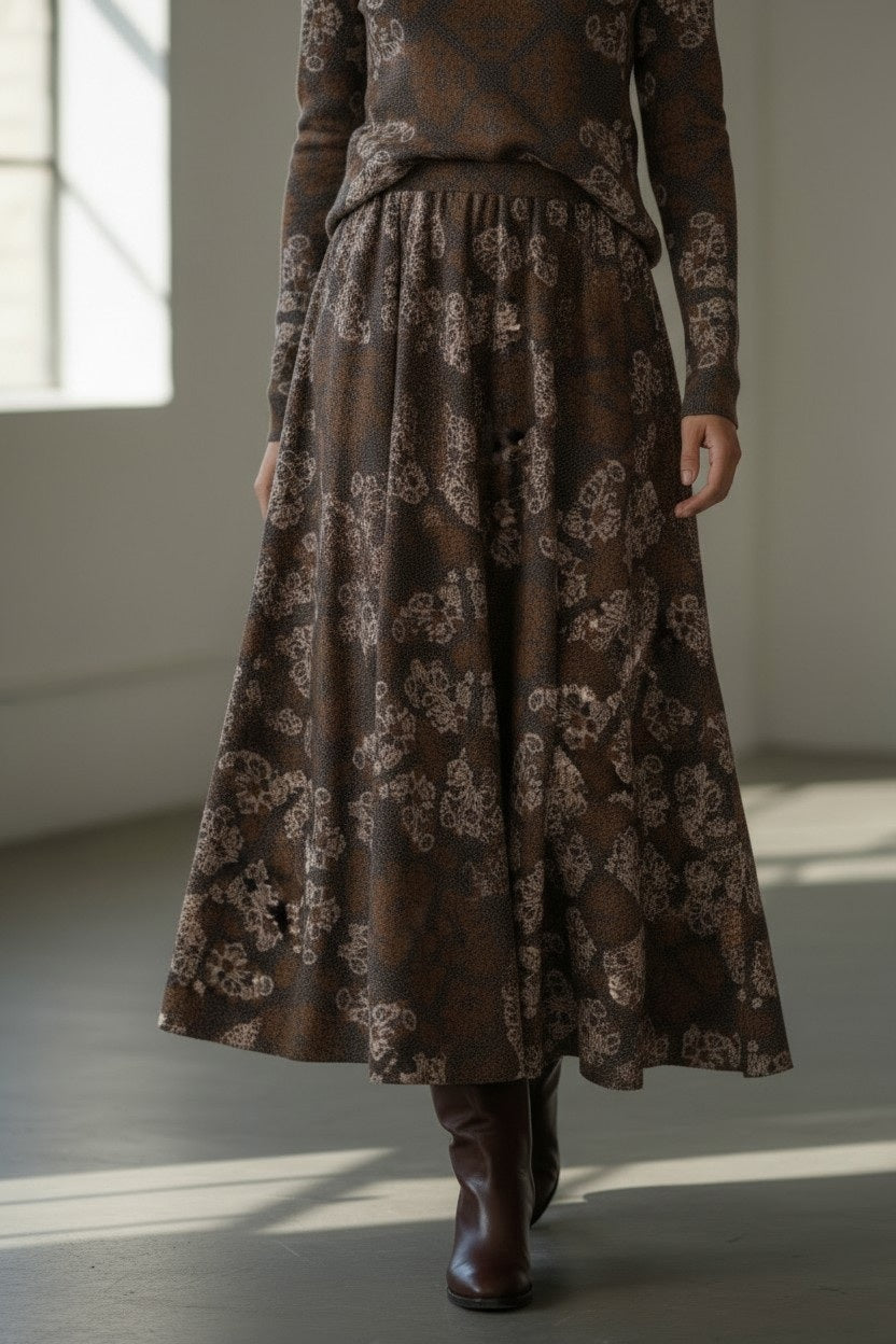 A woman stands indoors wearing the FRANCO FERRARI FRAGOLINA skirt, its floral motif flaring gracefully below a long-sleeved brown top, paired with brown boots. She smiles softly as sunlight streams through large windows in the background.