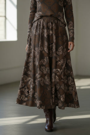 A woman stands indoors wearing the FRANCO FERRARI FRAGOLINA skirt, its floral motif flaring gracefully below a long-sleeved brown top, paired with brown boots. She smiles softly as sunlight streams through large windows in the background.