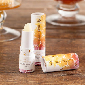 A tube of European Soaps, LLC Honey Lip Balm with its cap removed. The tube is mostly white with honeycomb and bee designs. The label text reads "lip balm, baume lèvres, .1 oz / 4 g." Infused with organic honey and royal jelly, the cap displays similar honeycomb and bee designs along with the brand's name.