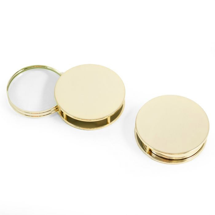 Two round, gold-plated BEYBERK INTERNATIONAL compacts are shown. One is closed; the other is open to reveal a 3X fold-out magnifier inside. Both have smooth, shiny surfaces and a simple, minimalistic design.