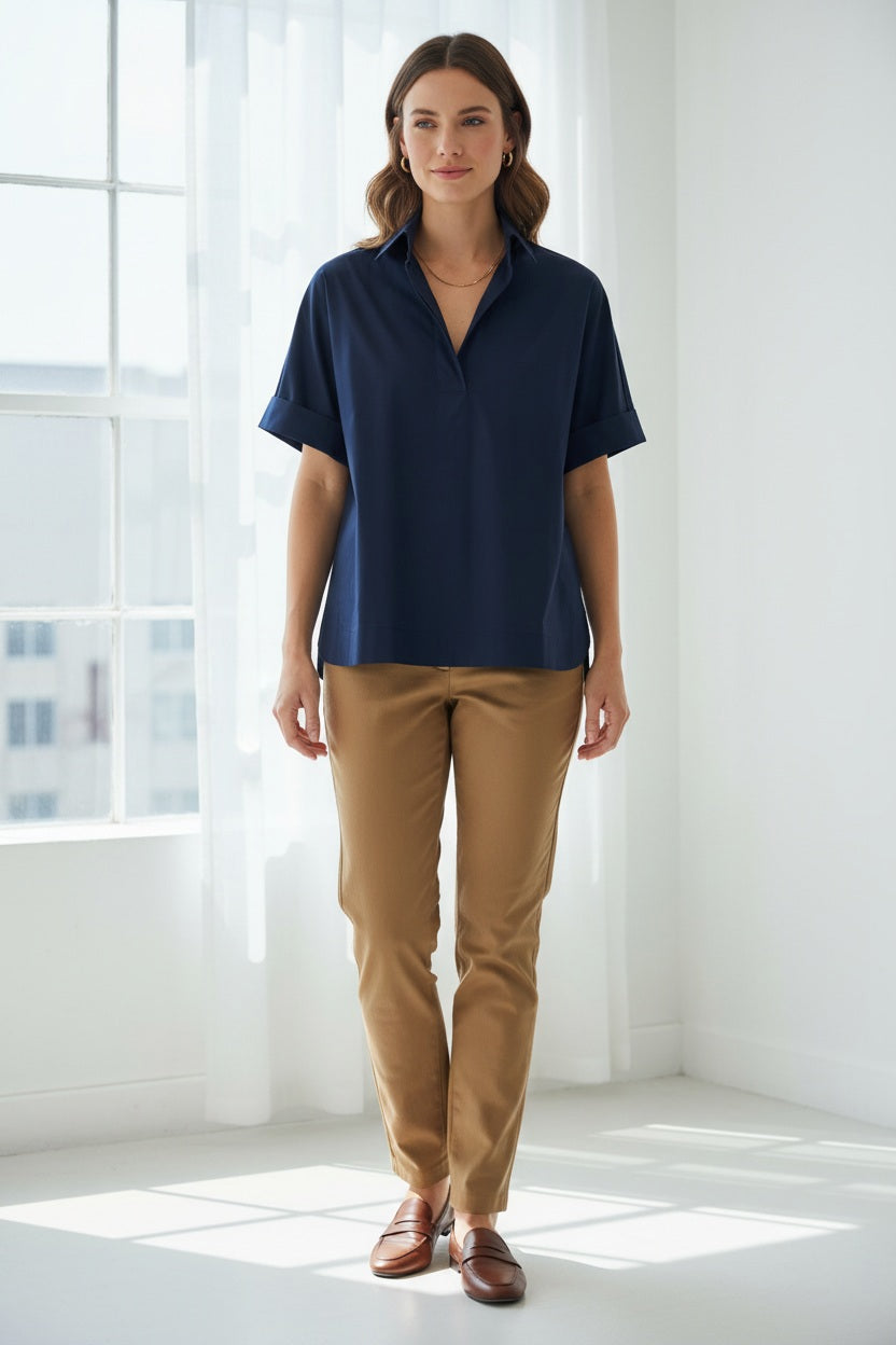 A woman with long brown hair is seen wearing the HINSON WU Cara Elbow Sleeve Shirt in Stretch Polished Cotton Navy—a loose-fitting, oversized short-sleeve shirt—paired with tan pants and gold jewelry, standing against a plain light gray background.