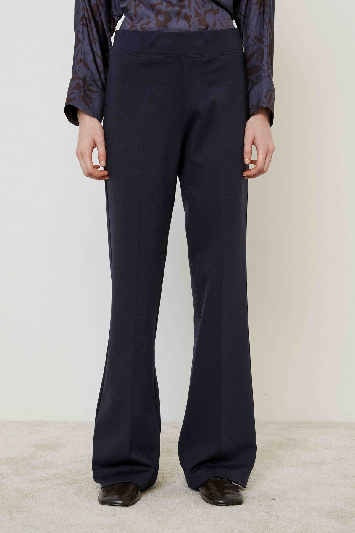 A person in MEIMEIJ PULL ON PONTE TROUSER PANT, a patterned long-sleeve blouse, and black shoes stands on a light carpet against a plain wall, highlighting this versatile MEIMEIJ wardrobe essential with a tailored look.