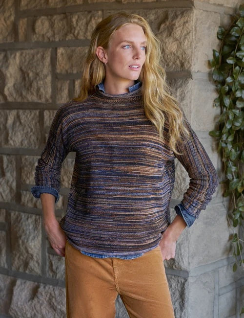 A woman stands near a window in a bright, minimal room, smiling and looking to the side while wearing the FRANK & EILEEN MONTEREY Rolled Funnel Neck Sweater in Pure Italian Cotton Harvest Stripes layered over a chambray shirt, paired with tan pants and brown ankle boots.