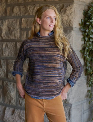 A woman stands near a window in a bright, minimal room, smiling and looking to the side while wearing the FRANK & EILEEN MONTEREY Rolled Funnel Neck Sweater in Pure Italian Cotton Harvest Stripes layered over a chambray shirt, paired with tan pants and brown ankle boots.