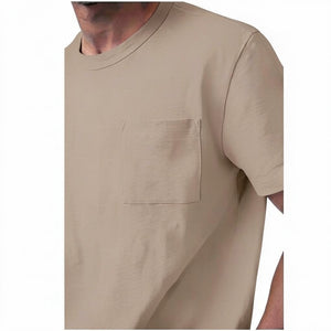 A man with short curly hair and a beard wears the MACK WELDON Heavyweight Slub Cotton Pocket T-Shirt in Desert Taupe and dark pants, standing against a plain white background.
