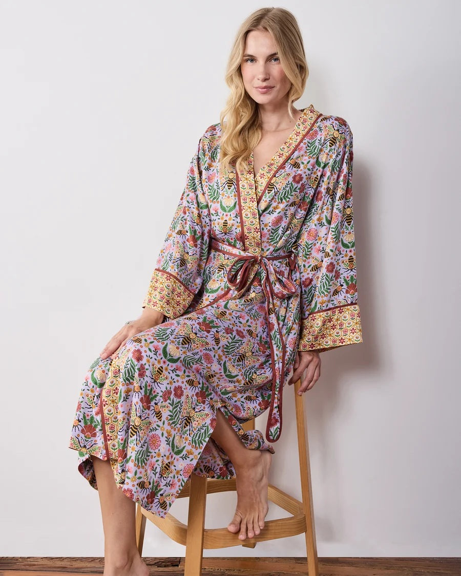 A woman wears a PRINTFRESH - BEE TAPESTRY ECO SATIN ROBE IN LAVENDER, featuring a lavender floral-print with a repeating pattern of bees, flowers, and vines with a contrasting yellow and red patterned trim.