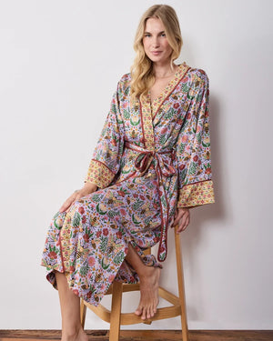 A woman wears a PRINTFRESH - BEE TAPESTRY ECO SATIN ROBE IN LAVENDER, featuring a lavender floral-print with a repeating pattern of bees, flowers, and vines with a contrasting yellow and red patterned trim.
