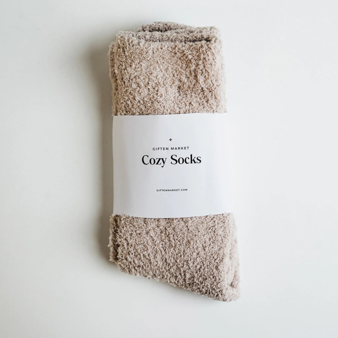 Beige fuzzy socks made from soft plush material are wrapped in a white &quot;Cozy Cloud Socks&quot; label displaying &quot;Giften Market&quot; and &quot;GIFTENMARKET.COM.&quot; These women&#39;s socks are neatly folded and viewed from above on a plain background.