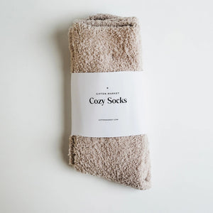 Beige fuzzy socks made from soft plush material are wrapped in a white "Cozy Cloud Socks" label displaying "Giften Market" and "GIFTENMARKET.COM." These women's socks are neatly folded and viewed from above on a plain background.