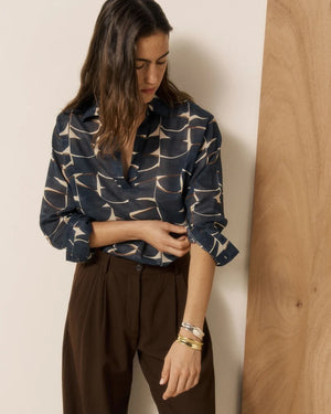 A woman stands in a bright room, smiling, wearing the HARTFORD - CHORUS POPOVER SHIRT IN DARK BLUE from HARTFORD—a dark printed cotton blouse that exudes French elegance—paired with light blue high-waisted jeans and tan ankle boots. White curtains and sunlight stream through a window on the left.