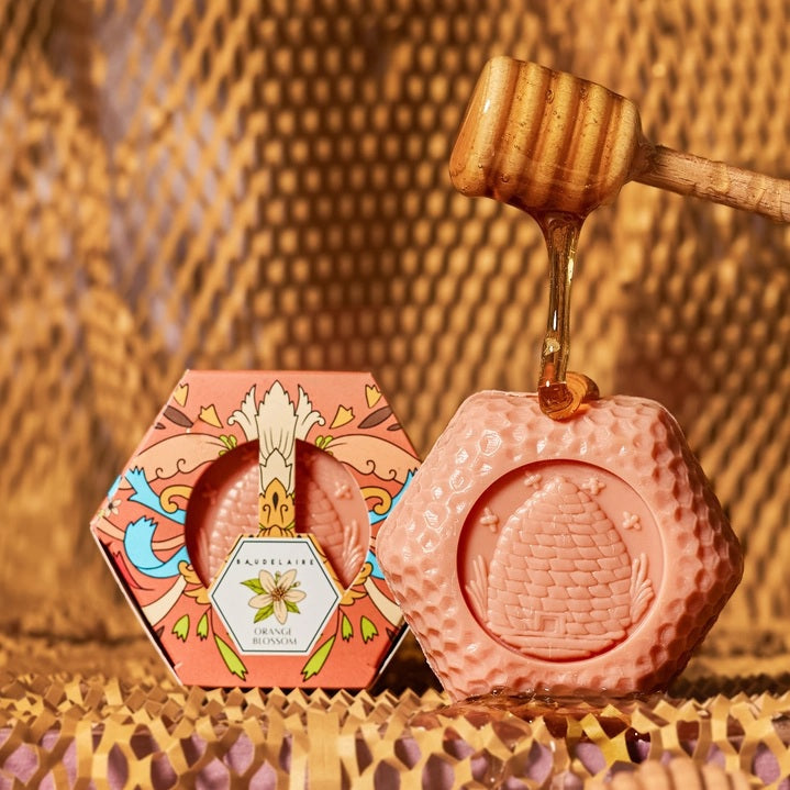 A hexagonal box of BAUDELAIRE - ORANGE BLOSSOM HONEY SOAP 3.5 OZ features a top panel displaying product benefits, a list of available scents—including Goat Milk Soap, Honey Soap, Royal Jelly, Lavender, Calendula, Pure Honey, and Orange Blossom—along with the ingredient list, barcode, and “Made in the USA” branding.