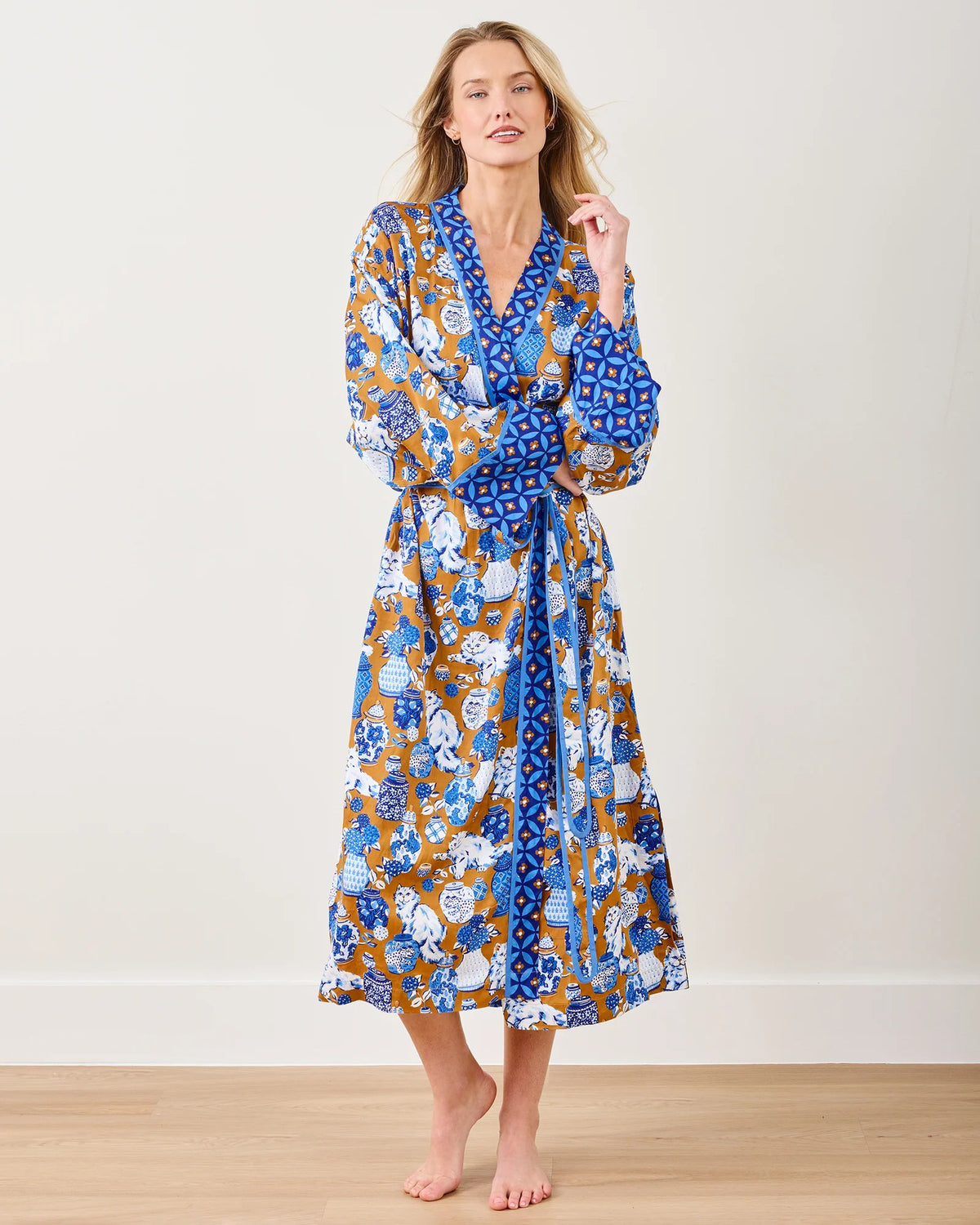 A person stands barefoot on a wooden floor, wearing the eco-friendly PRINTFRESH Satin Fancy Cat Robe with a blend of blue, white, and brown patterns. They pose with one hand on their chin and a neutral expression against a plain light-colored background.
