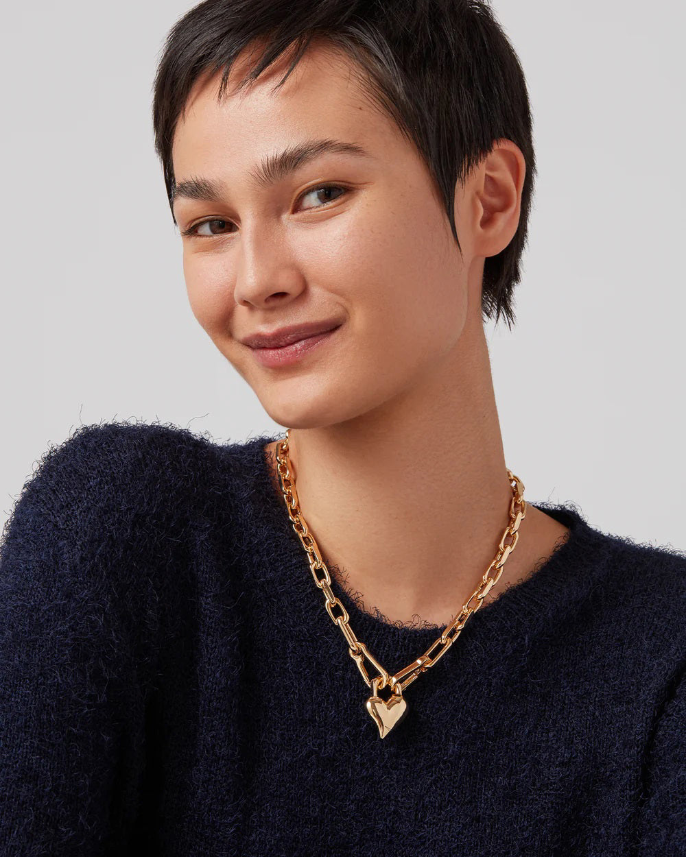 The JENNY BIRD HEART PENDANT NECKLACE showcases faceted gold chain links with a smooth, shiny heart charm at the center, elegantly set against a white background.
