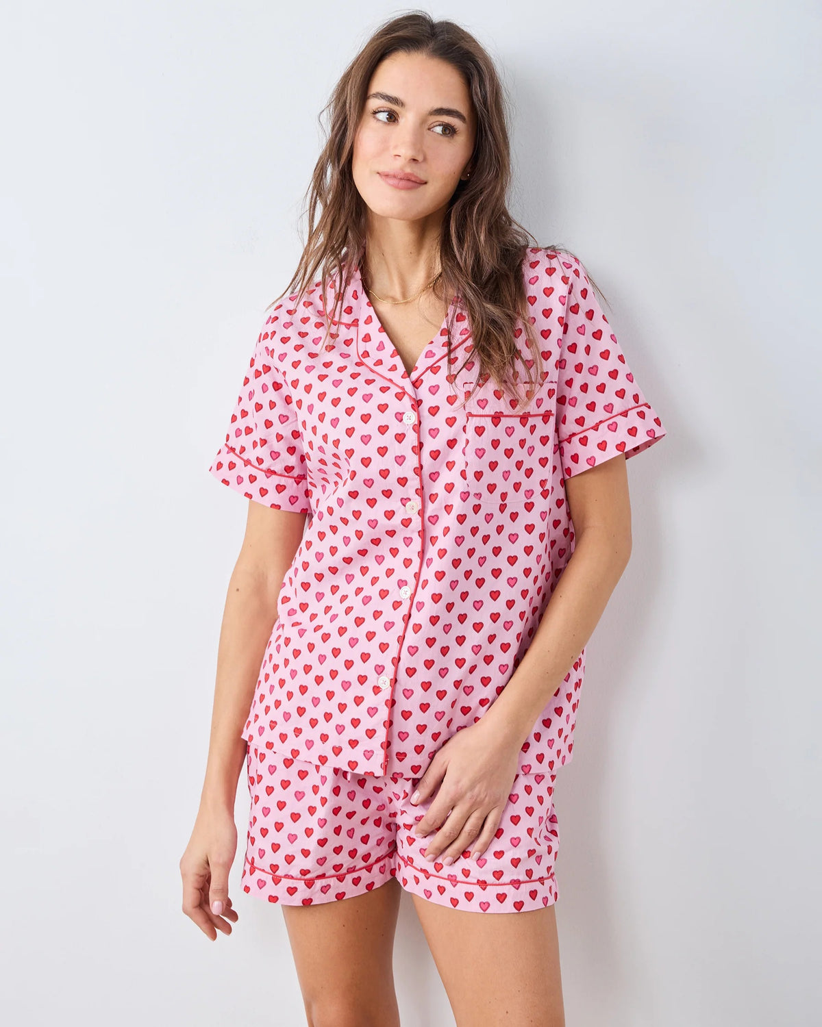 A person wearing the PRINTFRESH - MINI CANDY HEARTS PAJAMA SHORT SET by PRINTFRESH, featuring pink fabric with red heart patterns, stands with one hand in their shorts pocket. The image highlights the details of the pajama set and does not show the person's face.