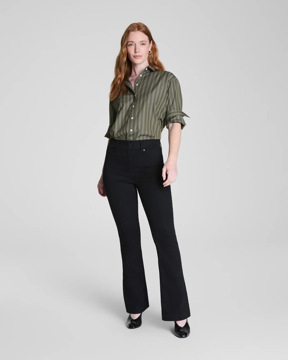 A woman stands indoors near a window, wearing a green button-up shirt styled with SPANX Pull On Flare Jean in Obsidian Rinse and black shoes. The background is minimal and gray.