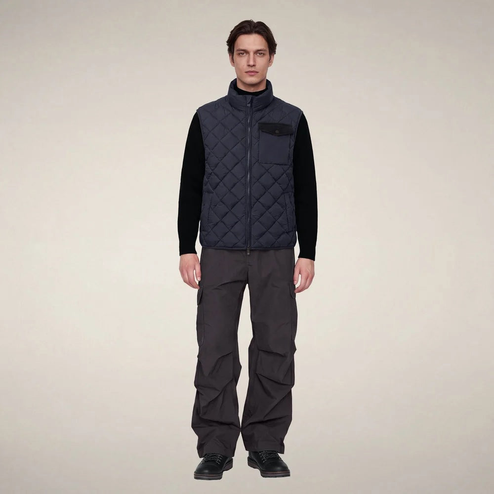 A person stands facing forward, wearing the SAVE THE DUCK - FABRICE Puffer Vest in Blue Black from SAVE THE DUCK, featuring recycled Plumtech insulation, layered over a black long-sleeve shirt and gray pants. They have a neutral facial expression and are posed against a plain white background.