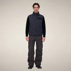 A person stands facing forward, wearing the SAVE THE DUCK - FABRICE Puffer Vest in Blue Black from SAVE THE DUCK, featuring recycled Plumtech insulation, layered over a black long-sleeve shirt and gray pants. They have a neutral facial expression and are posed against a plain white background.