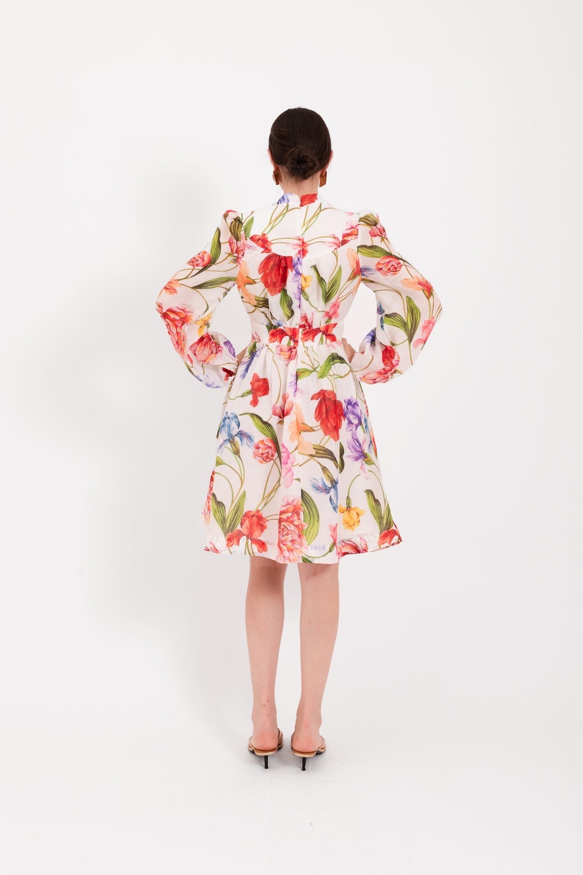 A woman stands against a white background, wearing the LAVI - CETERA FLORAL DRESS, a knee-length, long-sleeve dress by LAVI featuring a vibrant botanical print. She pairs it with open-toed sandals, her hair pulled back and her expression neutral.