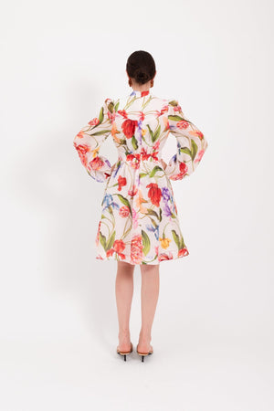 A woman stands against a white background, wearing the LAVI - CETERA FLORAL DRESS, a knee-length, long-sleeve dress by LAVI featuring a vibrant botanical print. She pairs it with open-toed sandals, her hair pulled back and her expression neutral.