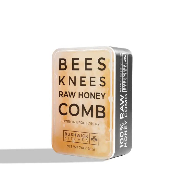 A rectangular tin container labeled "BUSHWICK KITCHEN - 'BEES KNEES' RAW HONEYCOMB" by Bushwick Kitchen. The off-white tin with black and orange text states "Born in Brooklyn, NY" and "Net Wt 7oz (198g)." One side is marked "100% Raw Honeycomb," containing pure, edible honey comb.