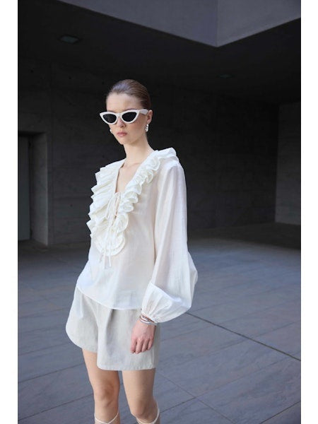 A woman in white sunglasses and white shorts poses against a dark modern building, wearing the ELEH - BLOUSE WITH LACES AND RUFFLES by ELEH—featuring cascading ruffles, sheer long sleeves, and lace-up white sandals. Her hair is tied back.