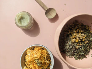 A small glass jar labeled "PINK HOUSE ORGANICS - CALM BALM" containing a light green, soothing balm. The jar is open, and the white lid is placed next to it on a neutral background. Ideal for all skin types, the balm appears smooth, indicating it hasn't been used yet.