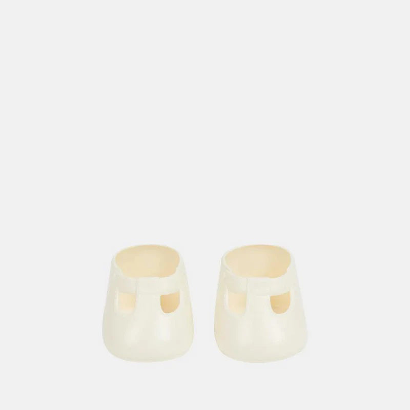 A close-up of a baby wearing OLLI ELLA USA DINKUM DOLL SHOES in Mallow Pink and white socks. The baby is seated on a soft, pale pink textured blanket, dressed in light brown clothing. Only the lower half of the baby's body is visible.