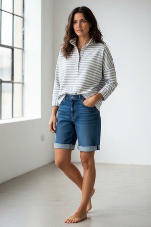 A person wearing sunglasses, denim shorts, and the MERSEA Emerson Pop Up Collar Top in Slate Blue Stripe stands on a rocky beach with buildings and trees in the background.