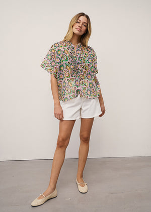 A woman with blonde hair poses casually against a plain background, wearing the 0039 ITALY DAISY BLOUSE IN LILAC AND GREEN PRINT, styled with white shorts and beige flats.