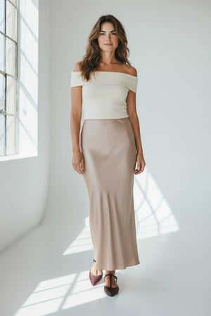 A person wearing the MILVA MI - LONG SATIN SKIRT IN SAND by MILVA, paired with pointed black shoes and standing against a plain, light background.