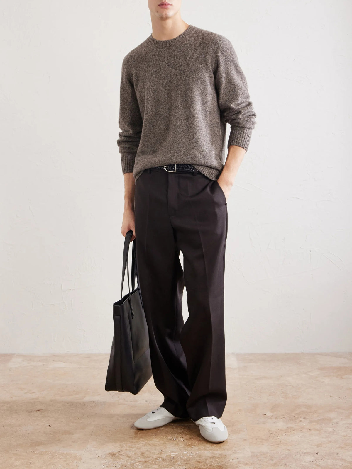 A person wearing the HARTFORD Seamless Wool Crew Pullover in Mushroom, an Italian-made sweater, paired with dark pants, is seated with one arm resting on their knee against a plain light background.