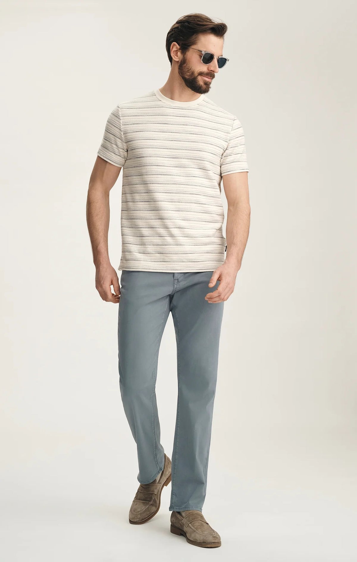 A man stands with his back to the camera against a plain light background, wearing a beige striped short-sleeve shirt, 34 HERITAGE Charisma Relaxed Straight Leg Pant in Flint Stone, brown suede shoes, and sunglasses.