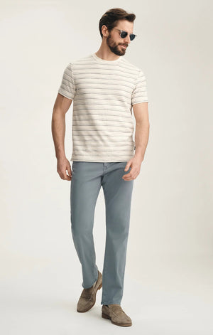 A man stands with his back to the camera against a plain light background, wearing a beige striped short-sleeve shirt, 34 HERITAGE Charisma Relaxed Straight Leg Pant in Flint Stone, brown suede shoes, and sunglasses.