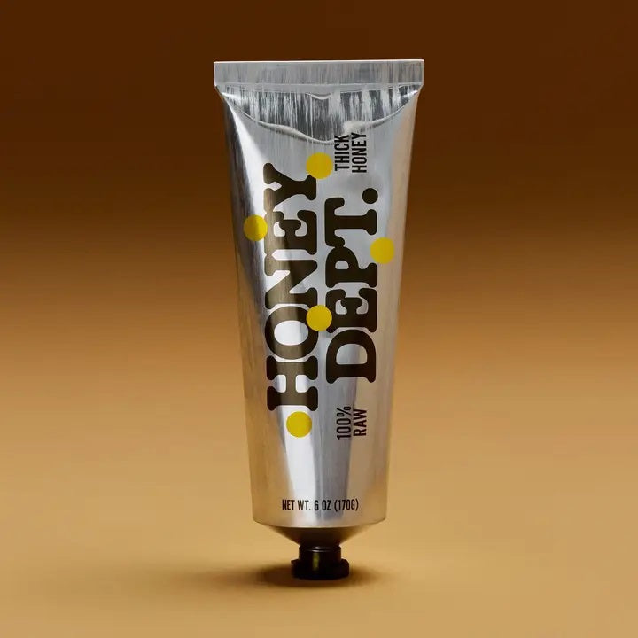 A metallic tube labeled “HONEY DEPARTMENT- THICK HONEY TUBE” stands upright against a brown gradient background. The design features yellow dots and bold black text, resembling a toothpaste or paint tube—making it perfect for dispensing thick honey with ease from HONEY DEPARTMENT.