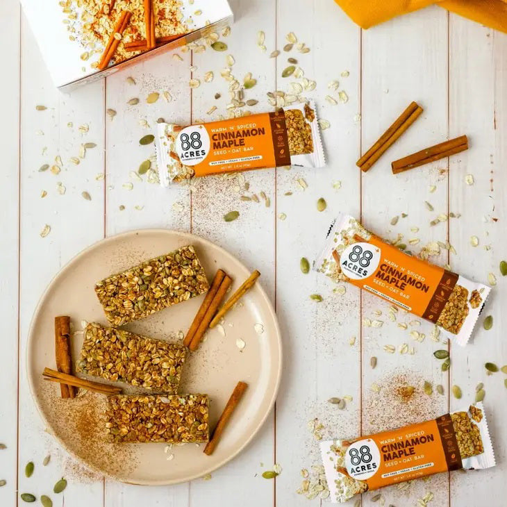 Image of an 88 ACRES Cinnamon Maple Seed + Oat Bar. The packaging is white with an orange section that displays the brand "88 ACRES" and the flavor details. The package showcases the nut-free, vegan, and gluten-free seed bar sprinkled with oats and seeds.