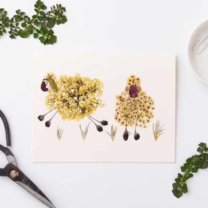 The PETAL PEOPLE PRESS Sheep Greeting Card by Petal People Press showcases a unique design, featuring two sheep artistically crafted from yellow dried flowers with tiny branches for legs and black flower heads as faces. Set against a white background, this charming scene evokes country life and is beautifully complemented by surrounding green leaves, a pen, and scissors.